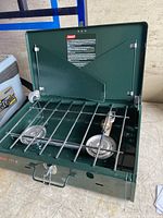 Green Coleman two-burner propane stove with lid open, showing burners and metal grates.