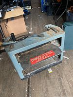 Front three-quarter view of benchtop planer showing infeed/outfeed rollers, cutter head warn label, carry handle and surface wear.