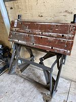 Folded wooden top of workbench showing multiple holes, scratches, and attached knobs. Metal foldable legs are visible below with some dirt around.