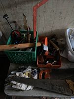Multiple hand saws, hammers, gloves, and clamps arranged on a table and inside a green crate and orange box.
