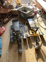 Two vintage NOMA GT Super snow racers placed side by side on a wooden floor with visible wear and various other items in the background.