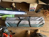 Portable hammock with stand folded and placed in a carrying bag on wooden floor, surrounded by various tools and equipment.