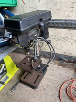 Photo showing black Jobmate drill press with base, adjustable table with clamp, power motor, and control lever.
