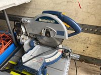 Clear side view of Mastercraft mitre saw placed on a workbench showing circular blade, adjustable angle base, handle and blade guard.
