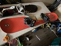 Bottom side of snowboard with orange base and attached black and white bindings, visible used condition.