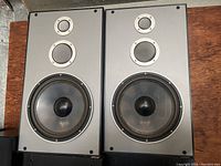 Pair of large 3-way tower speakers with visible woofer, midrange, and tweeter drivers in silver faceplates.