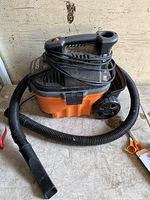 Orange and black Ridgid portable wet/dry vacuum with black flexible vacuum hose and narrow nozzle. Power cord wrapped around top handle.