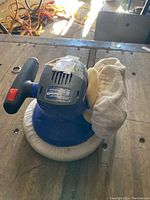 Blue Simoniz orbital polisher placed on a wooden surface. Shows black power switch on handle and polishing pads attached.