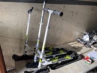Photo of three Stiga snow scooters with black skis, silver frames, and adjustable handlebars with black grips positioned indoors against a textured wall.