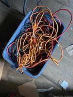 Top view of three coiled extension cords in a blue plastic tub on a concrete floor showing orange, black, and red cords tangled together.
