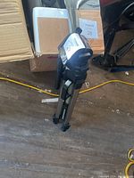 Folded silver-colored tripod leaning against boxes on floor, showing leg design and locking mechanisms with black plastic parts at joints and feet.