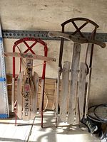 Two toboggans leaning against a wall showing full view, highlighting red and dark metal frames, wooden slats, and attached pulling ropes.