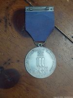 Front face of the silver-toned round medal attached to a purple ribbon with metal pin bar.