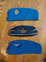 Overhead view of three military style caps: two blue Canadian Air Force caps with distinct badges and a black Scottish Rite Freemason cap with gold trim and silver eagle badge.