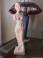 Front view of the cultured marble Venus De Milo statue showing detailed facial features, missing arms, and draped lower body standing on a square base with a lamp in the background.