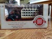 Front view of the diecast truck in original packaging showing green cab, red wheels and cream-colored cargo area with stacked crates and 100th anniversary logo.