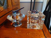 Two vintage table lighters side by side on wooden surface showing one flint lighter with circular base and glass top, and one clear glass square lighter with removable metal insert.