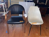 Two side chairs: one black leather wood framed chair and one white plastic chair with metal legs, displayed indoors on wood floor.