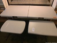 Three folding tables grouped together; largest folded in half on the floor, two smaller tables side by side on the ground, all white textured tops and black legs.