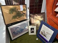Five framed Asian art pieces arranged on floor and leaning against objects. Includes prints and silk matted fabric art mainly depicting birds, plants, and architectural elements.