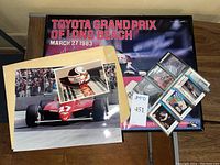 Wide shot showing the signed Toyota Grand Prix poster, several photographs of race cars, and trading cards laid out on a table.