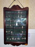 Full view of hanging wood display case with glass door containing 60 spoons
