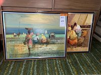 Pair of framed oil paintings, depicting Filipino coastal and village scenes, positioned on green patterned carpet, with artist signature visible on canvas.
