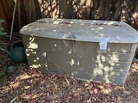 Large beige Rubbermaid resin outdoor storage bench with hinged lid, dirty with leaves and shadows on surface