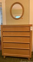 Front view of the light wood five drawer dresser with oval gold tone mirror hung above it