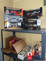 Overview of shelving unit showing six boxed 1:18 scale diecast race cars on top shelf and open box of smaller cars and cards below