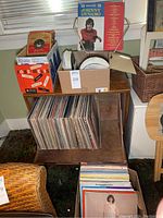 Wide view of lower shelf holding a row of twelve-inch vinyl LPs, upper surface with cardboard boxes of seven-inch records and loose sleeves, speaker behind.
