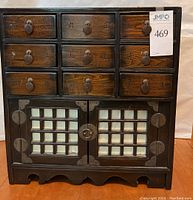 Front view of the wooden cabinet showing nine drawers with metal ring pulls and two doors with decorative metalwork and white grid panels.