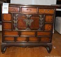 Front view showing central double doors with metal hinges and lock ring, flanked by multiple small drawers with brass handles, standing on carved legs.