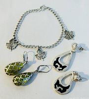 Silver chain bracelet with three maple leaf charms, two pairs of earrings laid out on white background