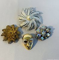 Top-down view of four vintage brooches: silver pinwheel with pearl, gold floral, mask face, and rhinestone cluster