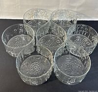 Seven clear glass Flora bowls arranged on dark background showing textured nature-inspired designs on sides and bottoms.