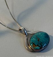 Close-up of the turquoise pendant showing the blue-green stone with brown veining set in sterling silver with a loop design and the 16-inch silver chain attached.