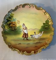 Close-up photo of the hand painted scene on the Royal Bayreuth plate showing the woman with geese and landscape background, highlighting the gold gilded detailed edge.
