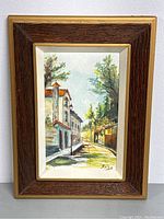 Full view of the framed painting showing street scene with buildings and trees, signed by artist.