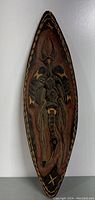 Full front view of elongated oval wooden shield with carved figure and painted patterns.