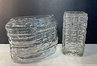 Photo of two clear art glass vases side by side on black background, showing rectangular textured geometric patterned vase, and irregular wavy shaped vase.