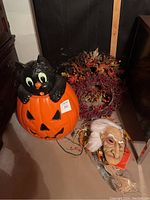 Photo showing large pumpkin decor with black cat, two wreaths with berries and foliage, and an old man Halloween mask with white hair and beard on the floor.