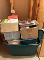 Green plastic storage bin full of various books and two cardboard boxes with more books inside.