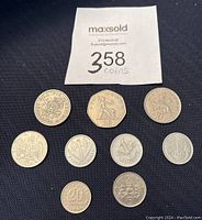 Top view of 10 coins on a dark fabric backdrop including UK 50 new pence, 10 new pence, Hungarian forint, French franc, Argentinian centavos, and others.