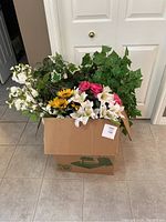 Box containing various artificial flowers and plants including white lilies, pink peonies, yellow sunflowers, and green ivy leaves.
