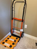 Full view of orange and black hand dolly standing upright against wall on carpet