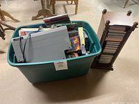 Wide shot showing green plastic bin filled with VHS tapes, DVDs, and an Emerson VHS player on top. A wooden vertical CD shelf is visible nearby containing multiple CDs.