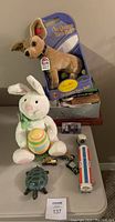 Overview photo showing Taco Bell dog plush in packaging, plush Easter bunny, toy vehicles including Esso tanker truck, turtle, small vintage cars, and various game boxes beneath.