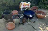 Overview of multiple clay, ceramic, and metal garden pots and garden decor items placed outdoors on concrete.