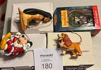 Four Christmas ornaments displayed on flat surface, including Pocahontas Meeko in box, brown dog with snowman, Santa Claus figure, and wooden rocking horse.